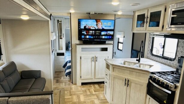 Interior compacto de autocaravana con kitchenette, televisor montado en la pared y zona de estar con sofá.