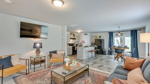 Un salon moderne avec un canapé, des fauteuils, une table basse et un téléviseur. On aperçoit une cuisine et un coin repas à l’arrière-plan.