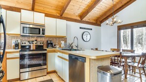 Cucina con forno a microonde, forno e lavastoviglie in acciaio inossidabile, pensili in legno, una sveglia e una finestra con vista sugli alberi.