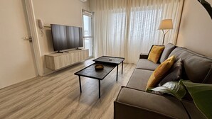 Un salon moderne avec un téléviseur à écran plat fixé sur une console en bois, une table basse de couleur sombre, un canapé avec des coussins et un lampadaire.