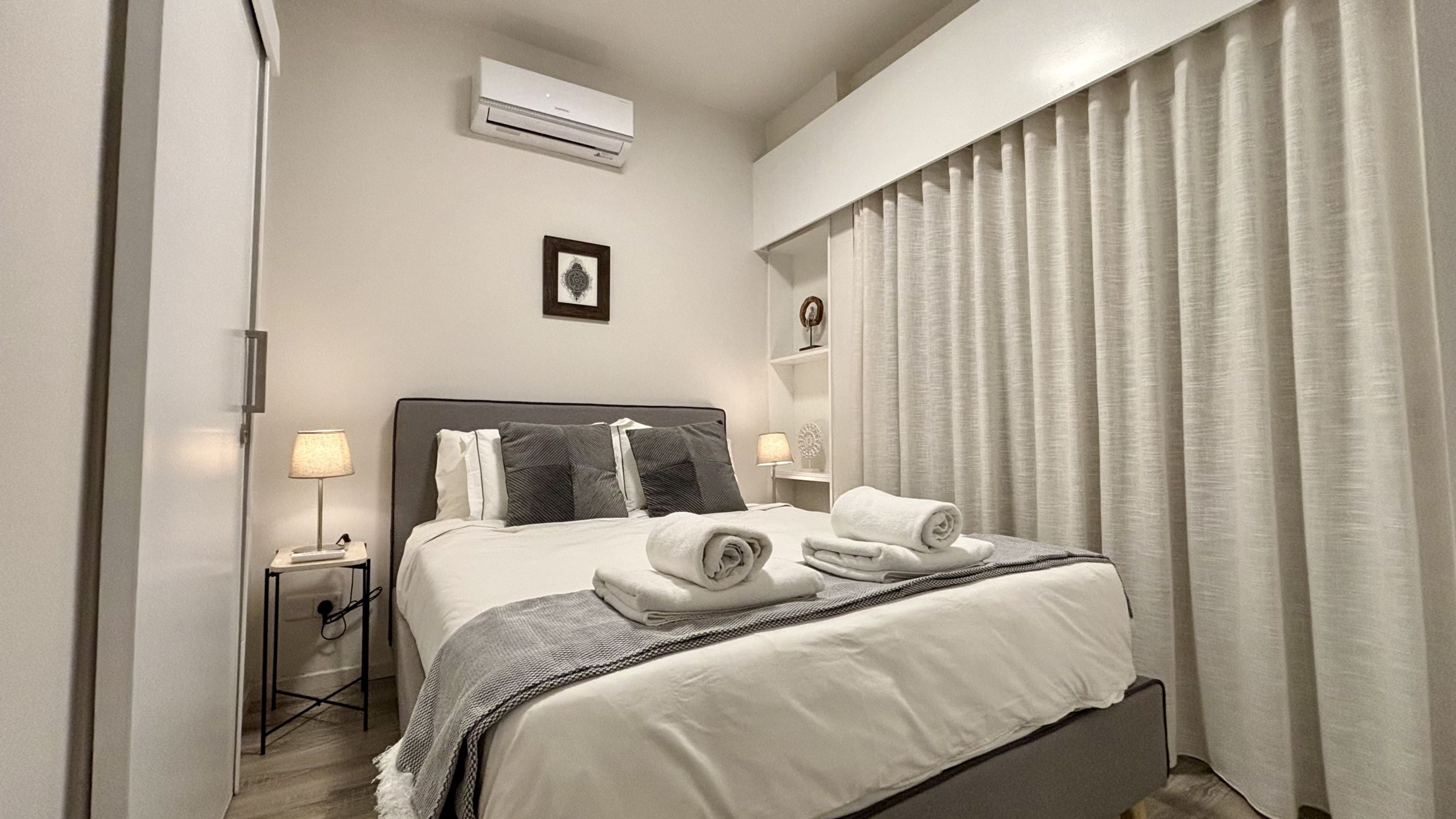 A hotel room with a bed, a bedside table with a lamp, an air conditioning unit, a framed picture on the wall, and a window with curtains.