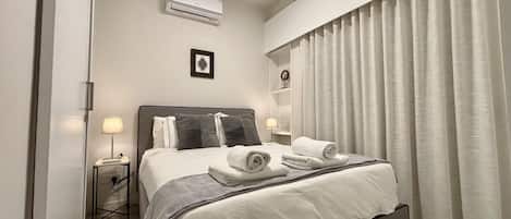 A hotel room with a bed, a bedside table with a lamp, an air conditioning unit, a framed picture on the wall, and a window with curtains.
