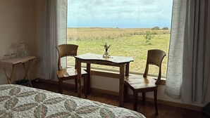 A bedroom with a large bed, a wooden table with two chairs, a small table with a pitcher and glasses, and a view of a grassy field through a window. - Simple yet enchanting abode  (Naalehu)