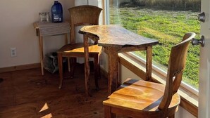 A room with a wooden table and chairs, a window with a view of a grassy field, and a door. - Simple yet enchanting abode  (Naalehu)