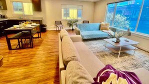 A modern living room with a sofa, a dining table, and a large window offering a view of the city. - The Seattle Haven - Modern Suites (Seattle)