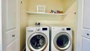 A laundry room with two front-loading washing machines and a shelf above them. - The Seattle Haven - Modern Suites (Seattle)