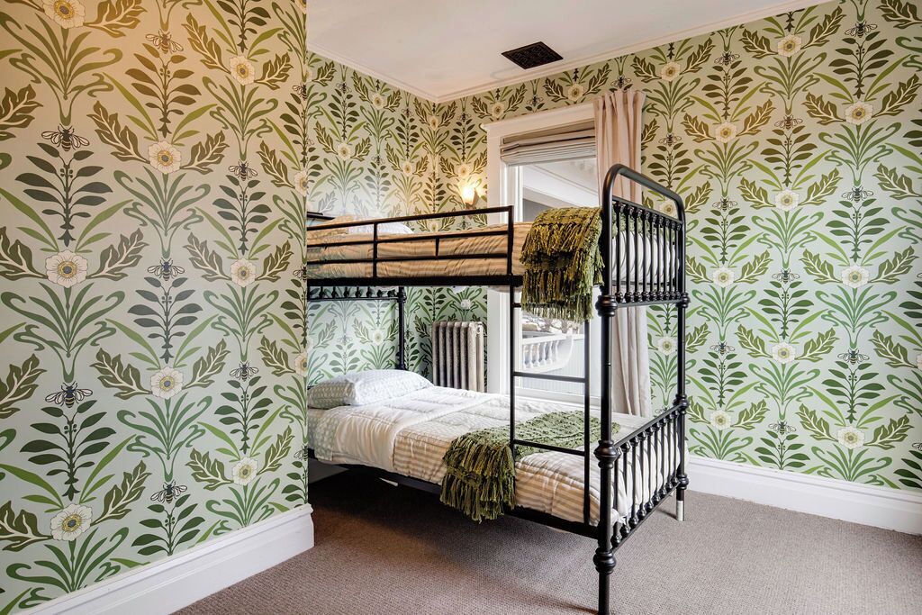 Een stapelbed in een kamer met bloemenbehang, een raam met gordijnen, en een radiator.