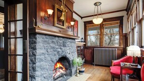 A stone fireplace with a fire, wooden paneling, a red chair, and a window with a view of trees. - Rosedale at Historic Hopffgarten House (Boise)