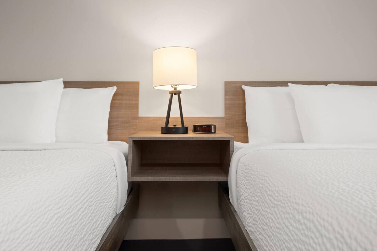A hotel room with two beds, a nightstand with a lamp, and a small table.