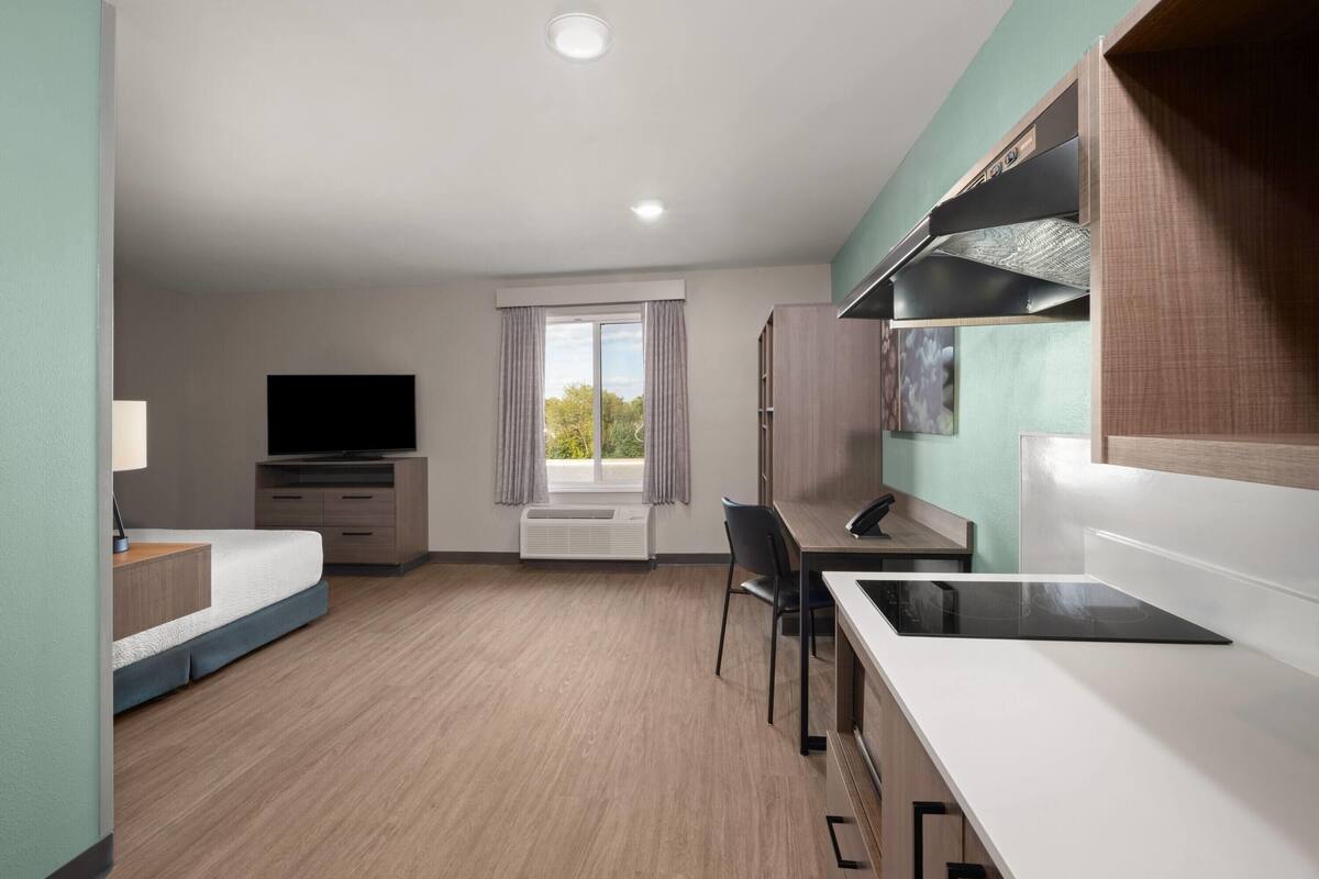A modern hotel room with a bed, desk, chair, TV, and kitchenette.