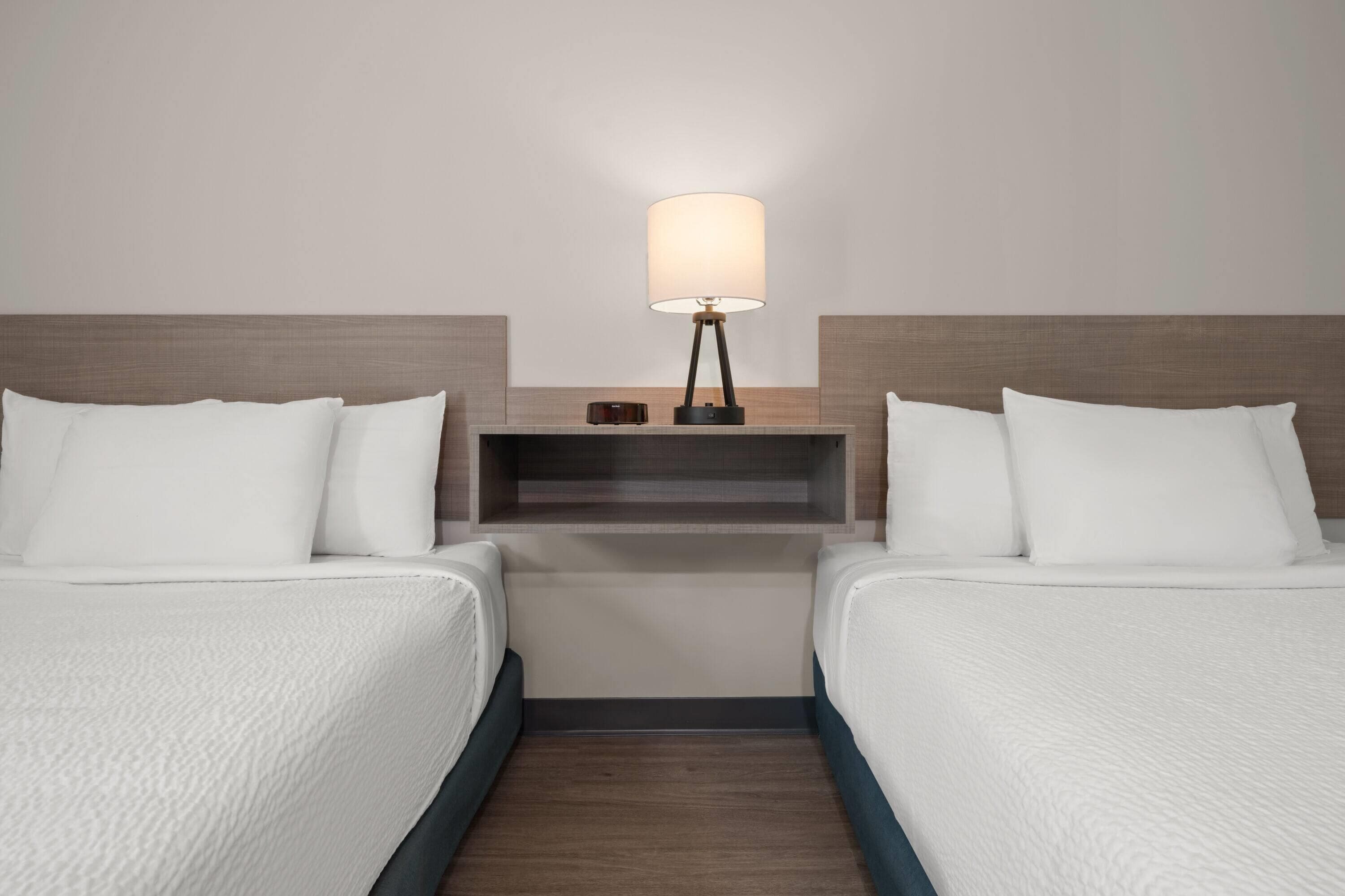 A hotel room with two beds, a nightstand with a lamp, and a shelf with a clock.