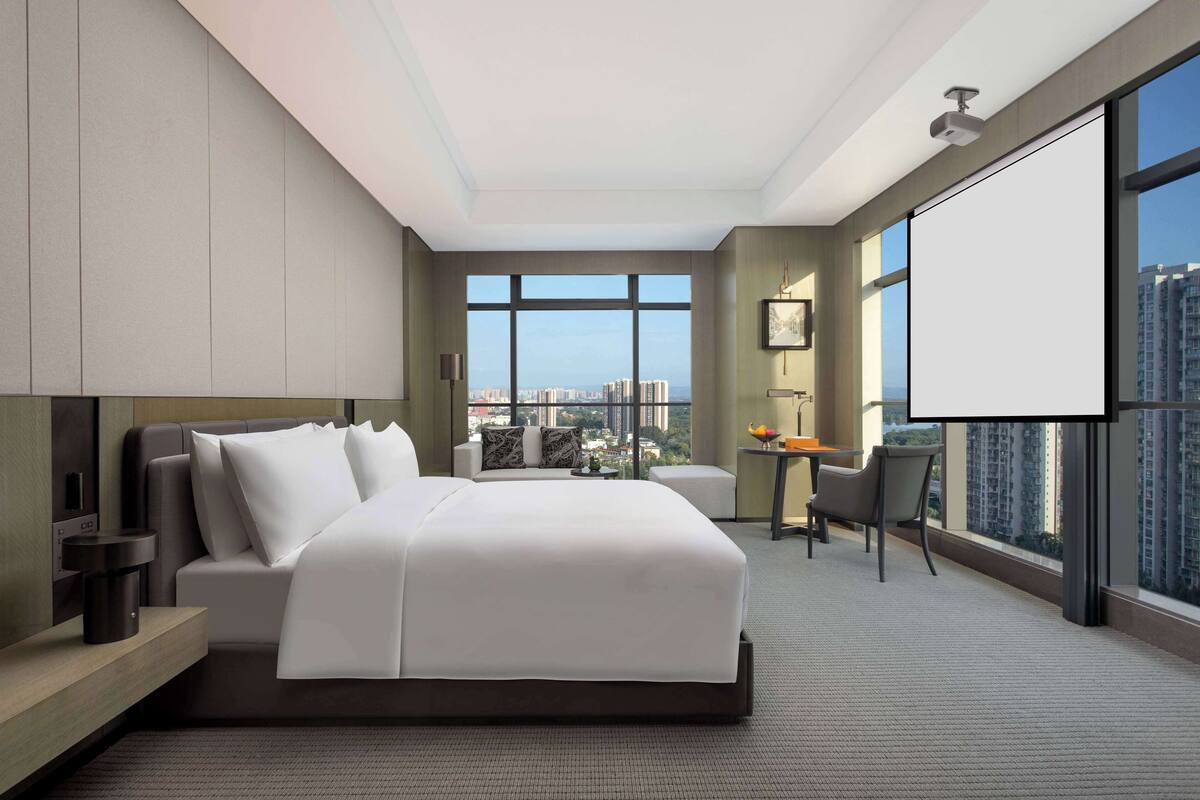 A modern hotel room with a large bed, a desk, and a view of the city through large windows.