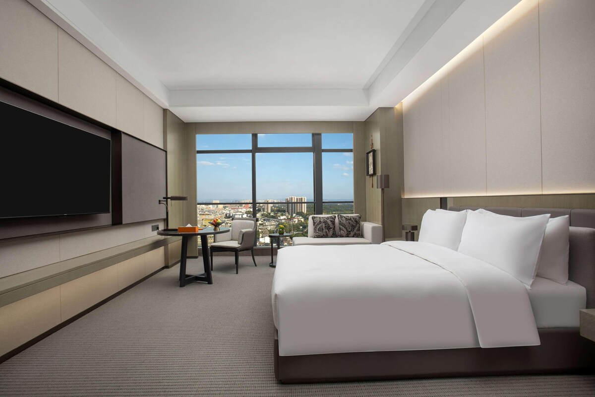 A hotel room with a large bed, a desk with a chair, a TV, and a view of a cityscape through large windows.
