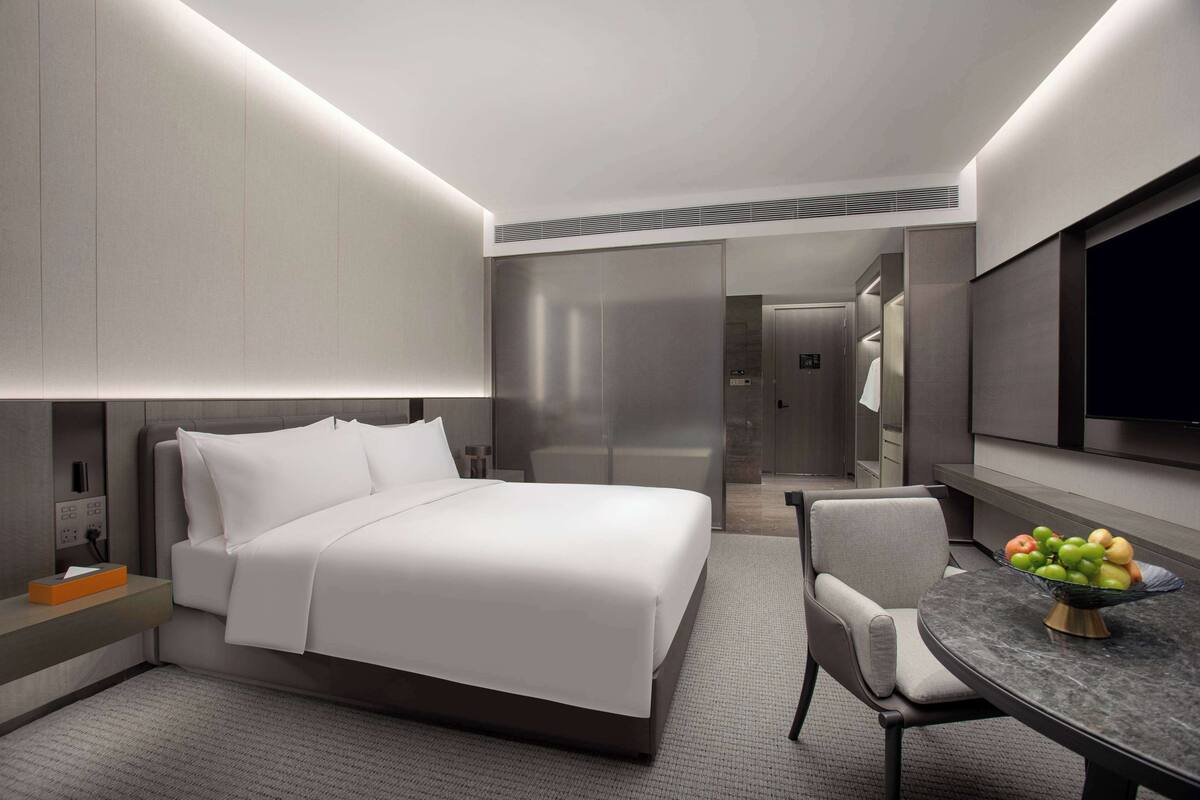 A modern hotel room with a large bed, a desk with a fruit bowl, a chair, and a TV.