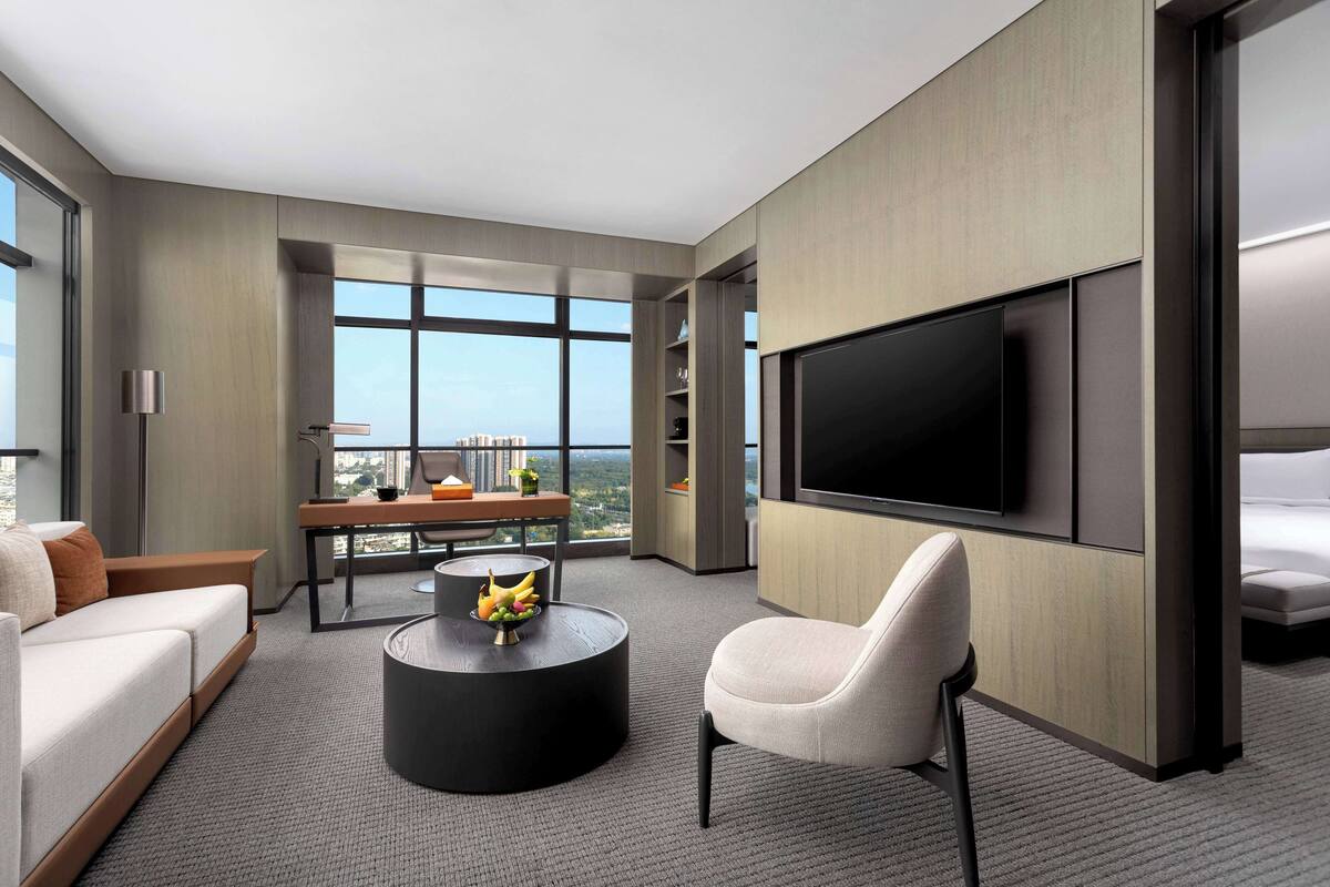 A modern hotel room with a large TV, a sofa, a dining table with a fruit bowl, and a city view.