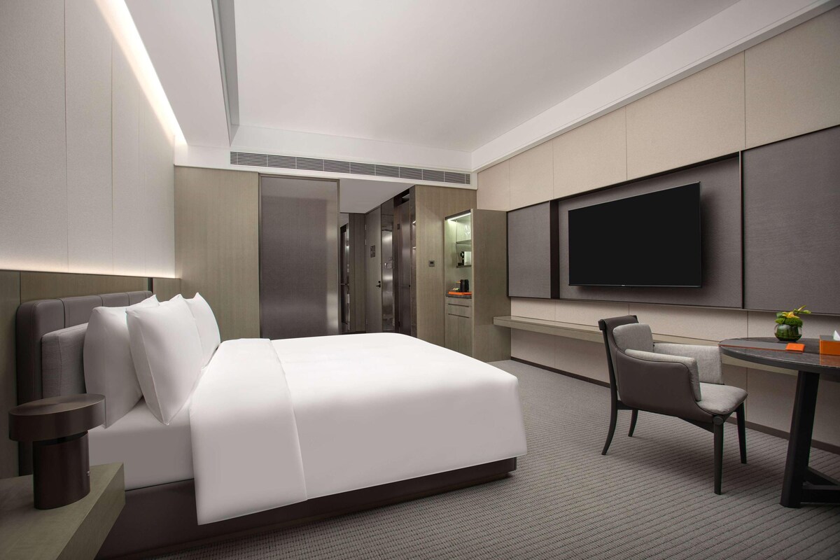 A modern hotel room with a large bed, a desk with a chair, a flat-screen TV, and a small table with a plant.