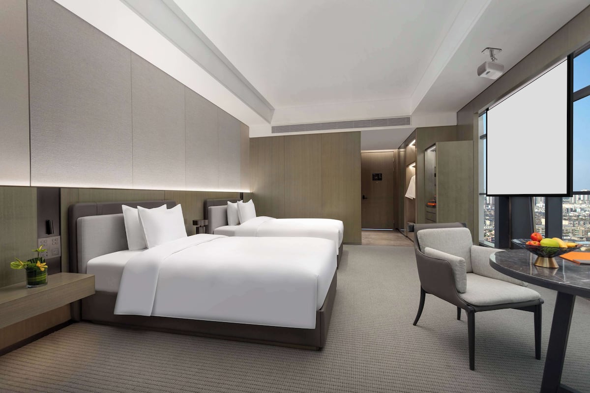 A modern hotel room with two beds, a desk, and a chair. There is a large window providing a city view.