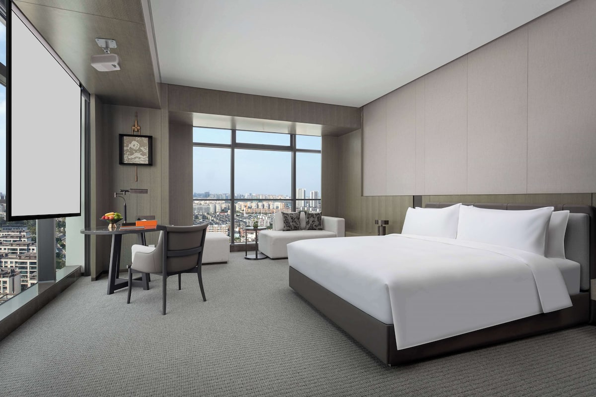A modern hotel room with a large bed, a desk with a chair, a small sofa, and a large window offering a city view.