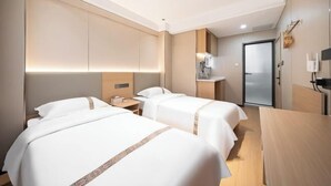 Habitación de hotel con dos camas, un escritorio, una silla, una mesita, una mesita de noche, una lámpara y una puerta.