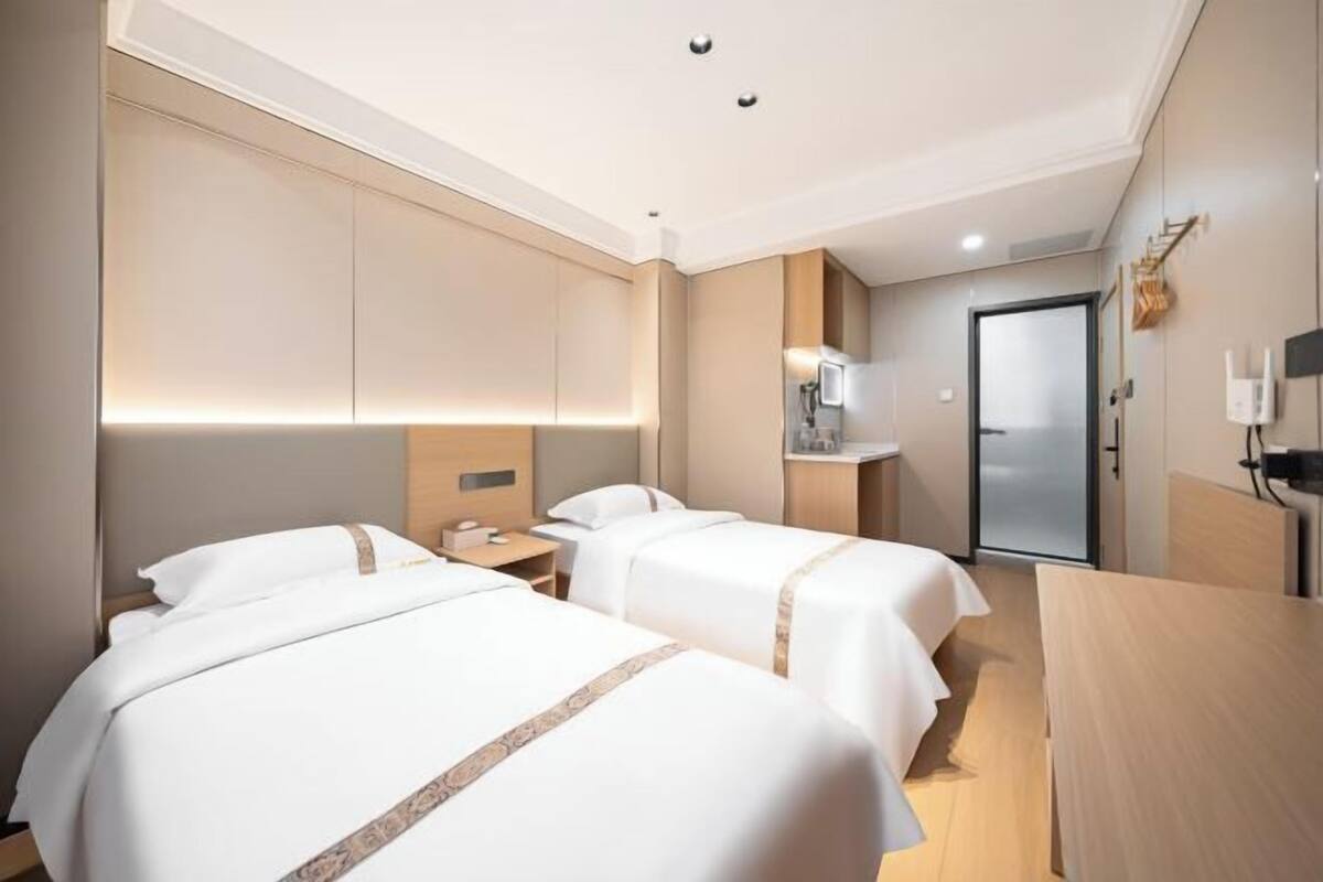 A hotel room with two beds, a desk, a chair, a small table, a nightstand, a lamp, and a door.