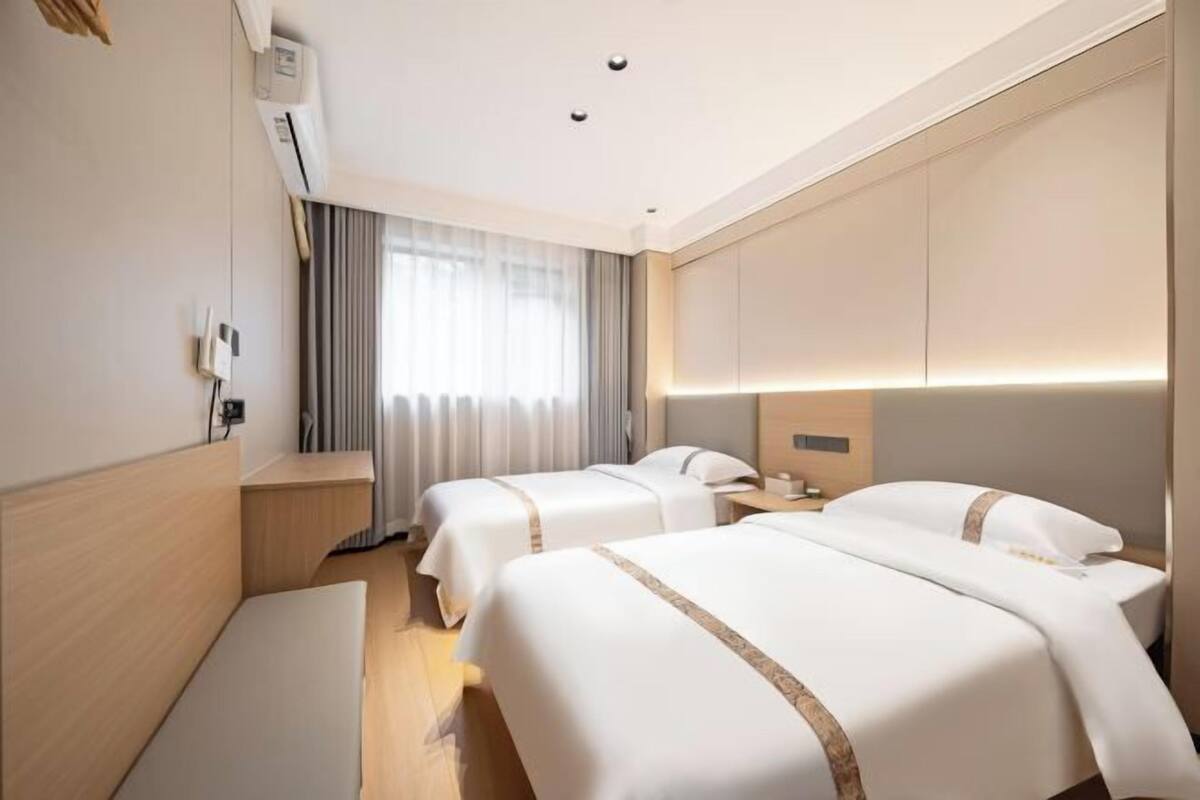 A hotel room with two beds, a desk, a chair, a window with curtains, and a wall-mounted air conditioner.