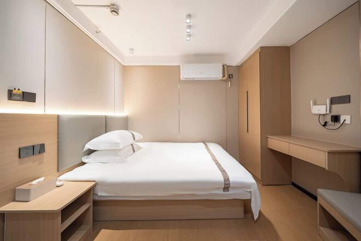A modern hotel room with a bed, bedside table, built-in wardrobe, and a desk area.