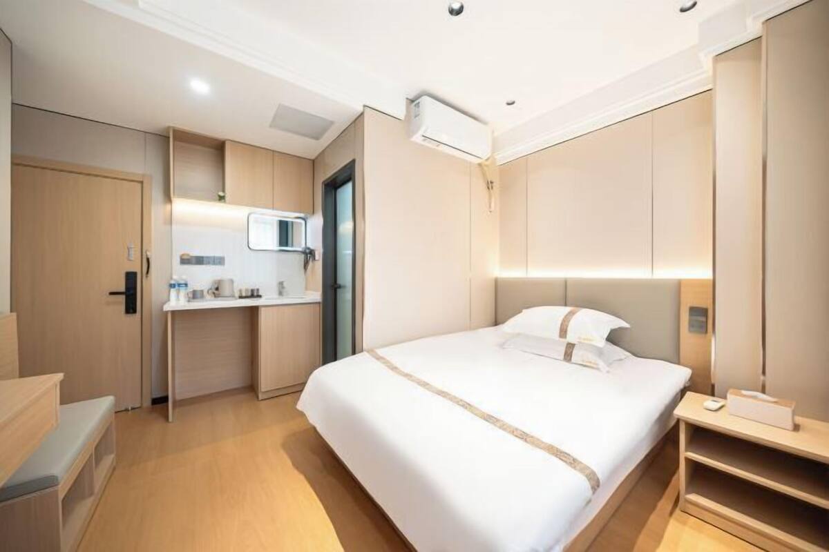 A modern hotel room with a bed, bedside table, built-in wardrobe, and a kitchenette area.