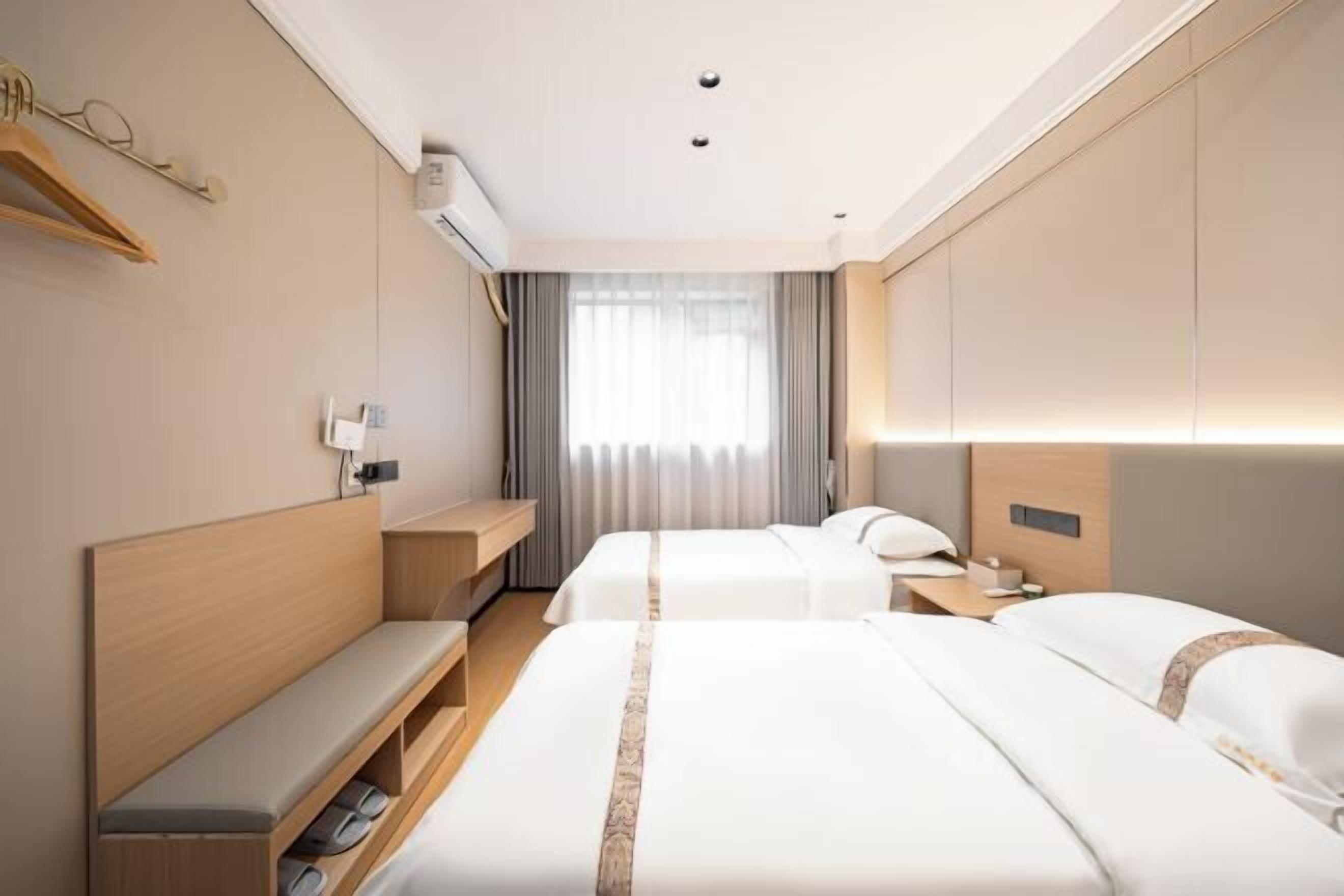 A hotel room with two beds, a bench, a bedside table, a nightstand, and a window with curtains.