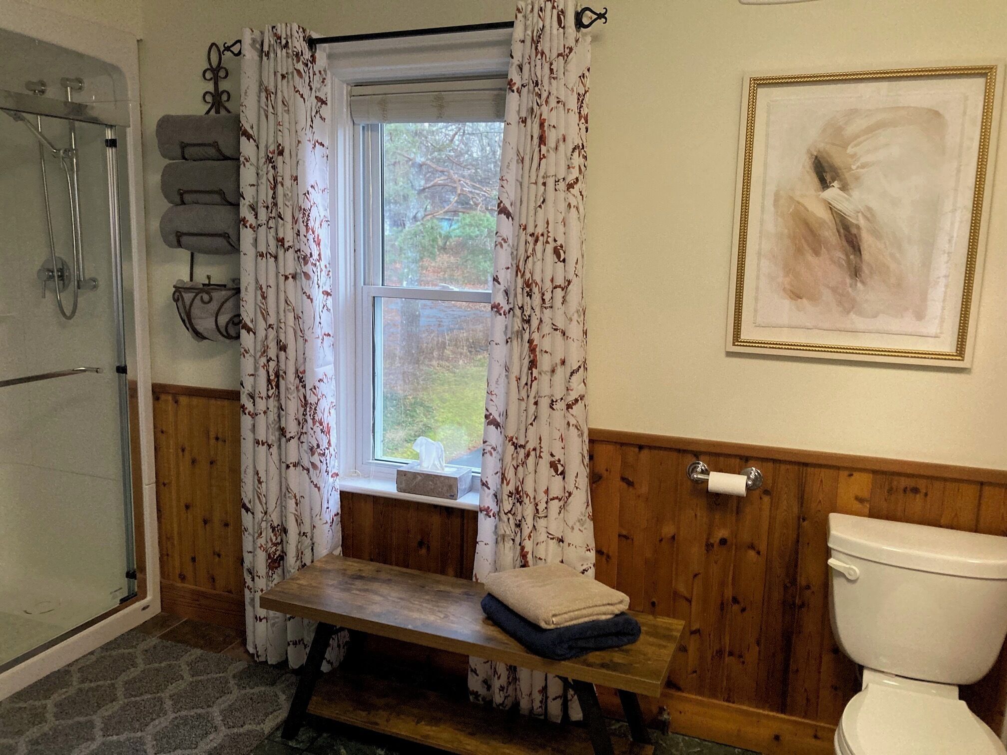 Un baño con ducha, una ventana con cortinas, un banco de madera, un inodoro y un cuadro enmarcado en la pared.