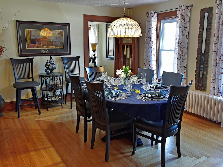 Une salle à manger avec une table ronde dressée pour six personnes, entourée de six chaises. Un lustre est suspendu au-dessus de la table, et un grand tableau encadré orne le mur.