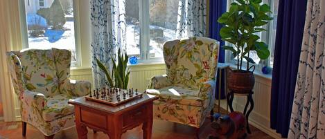 Un salon avec des fauteuils à motifs floraux, une table d’échecs et une plante en pot près d’une fenêtre ornée de rideaux.