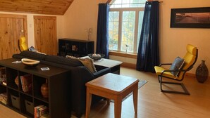 Una sala de estar acogedora con techo de madera, un ventanal con cortinas, un sofá negro, un sillón amarillo, una mesa de centro de madera y una estantería llena de libros y objetos decorativos.