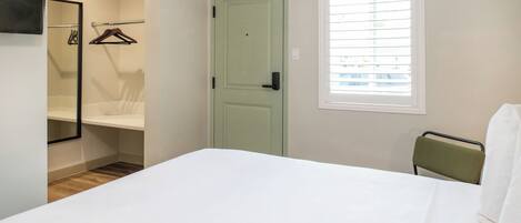 A bedroom with a bed, a green door, a window with blinds, a mirror, and a chair.