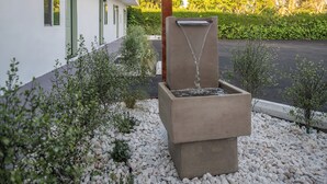 A modern outdoor water feature with a concrete structure, surrounded by white pebbles and small shrubs. - The LEEF Hotel (Glendale)