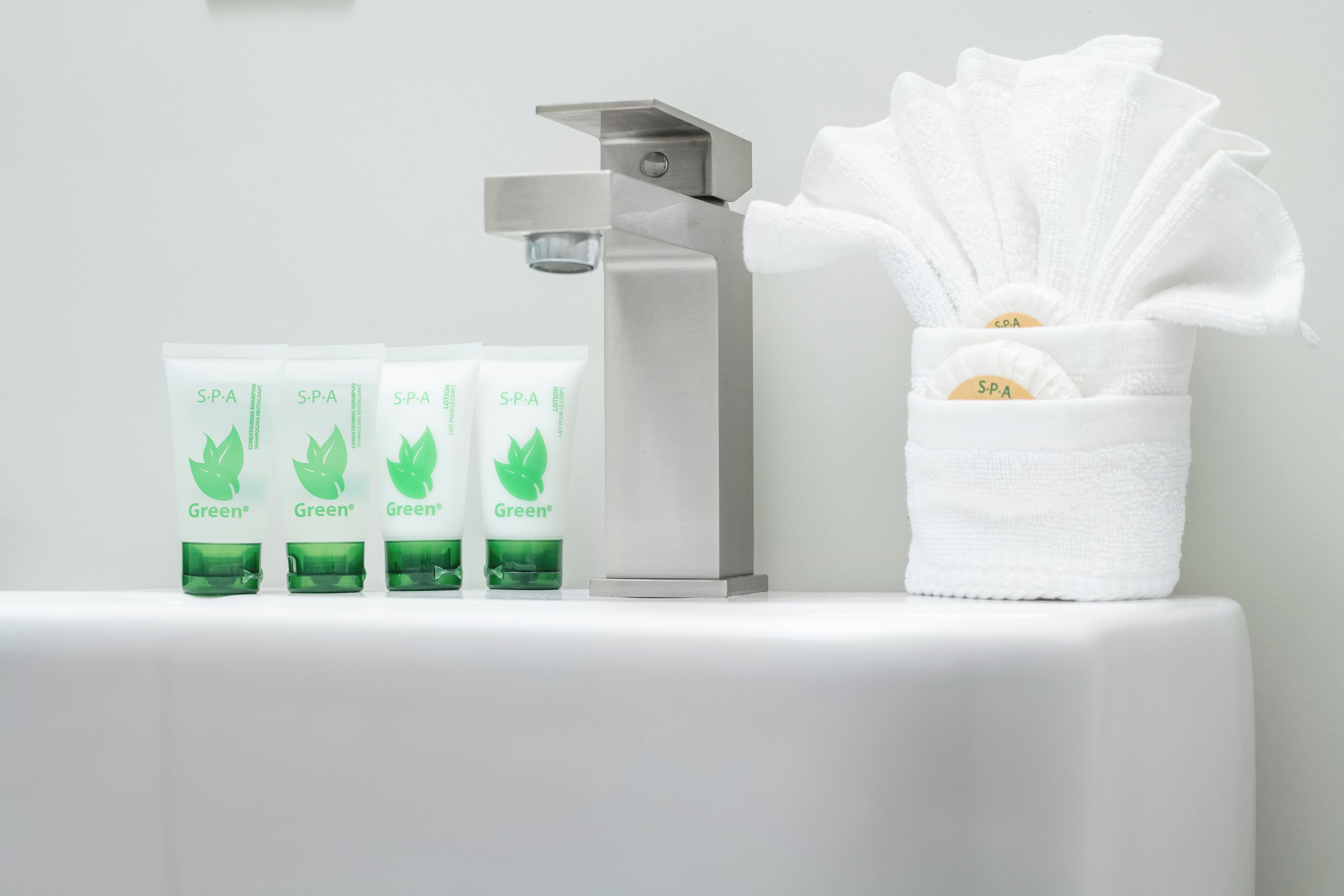 A bathroom with a white sink, a silver faucet, and a tissue holder with a white towel. There are four green bottles labeled "S.P.A Green" on the counter.