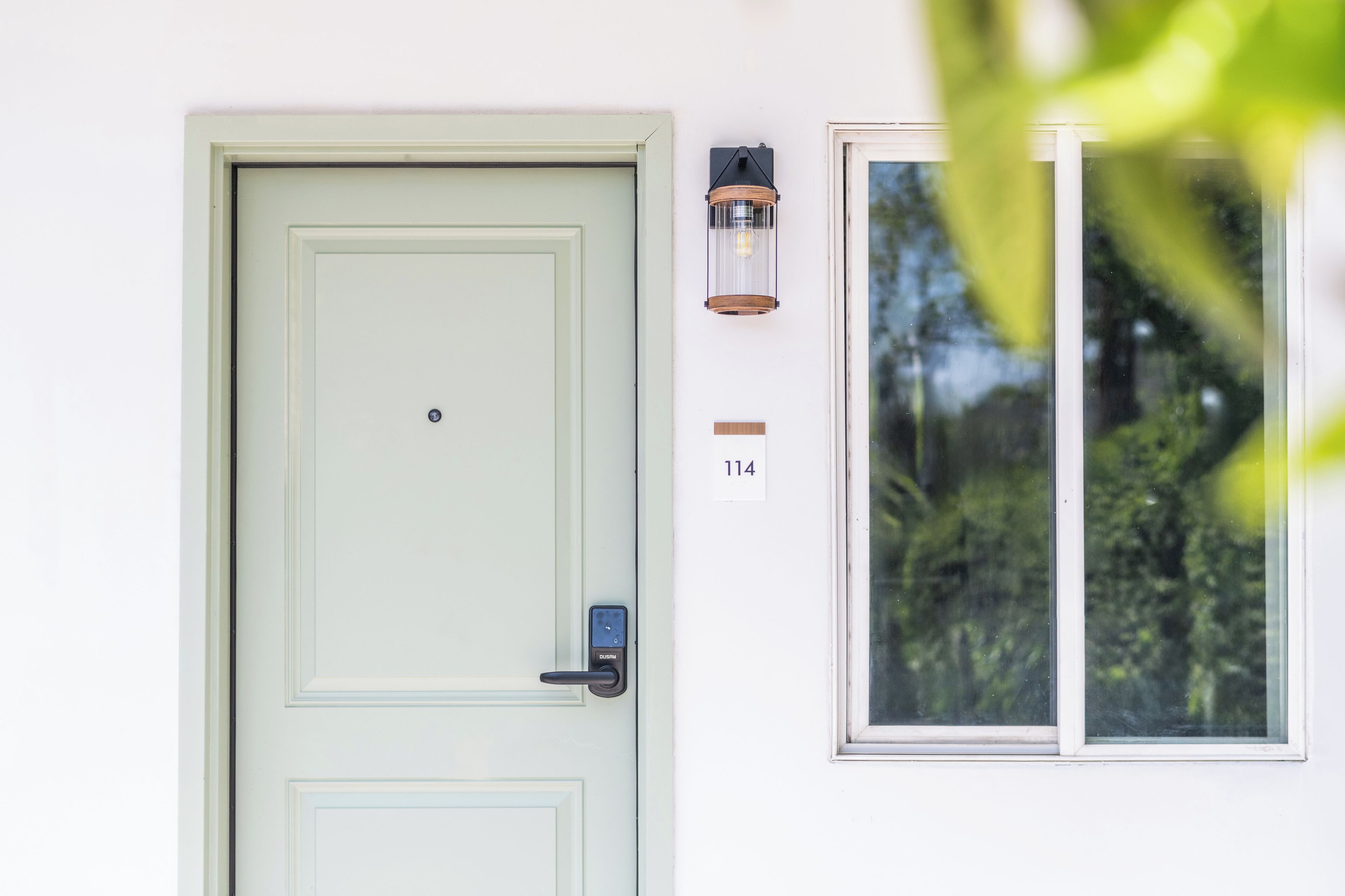 A light green door with a keypad lock, a wall-mounted number plate labeled "114", and a window with a view of greenery.