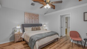 A bedroom with a bed, nightstands, a desk, a chair, a ceiling fan, and a door.