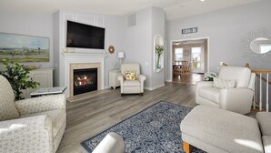 A living room with a fireplace, a television, and a staircase leading to another room. - Bayfront Coastal Paradise | Pool | Beach Access (Bethany Beach)