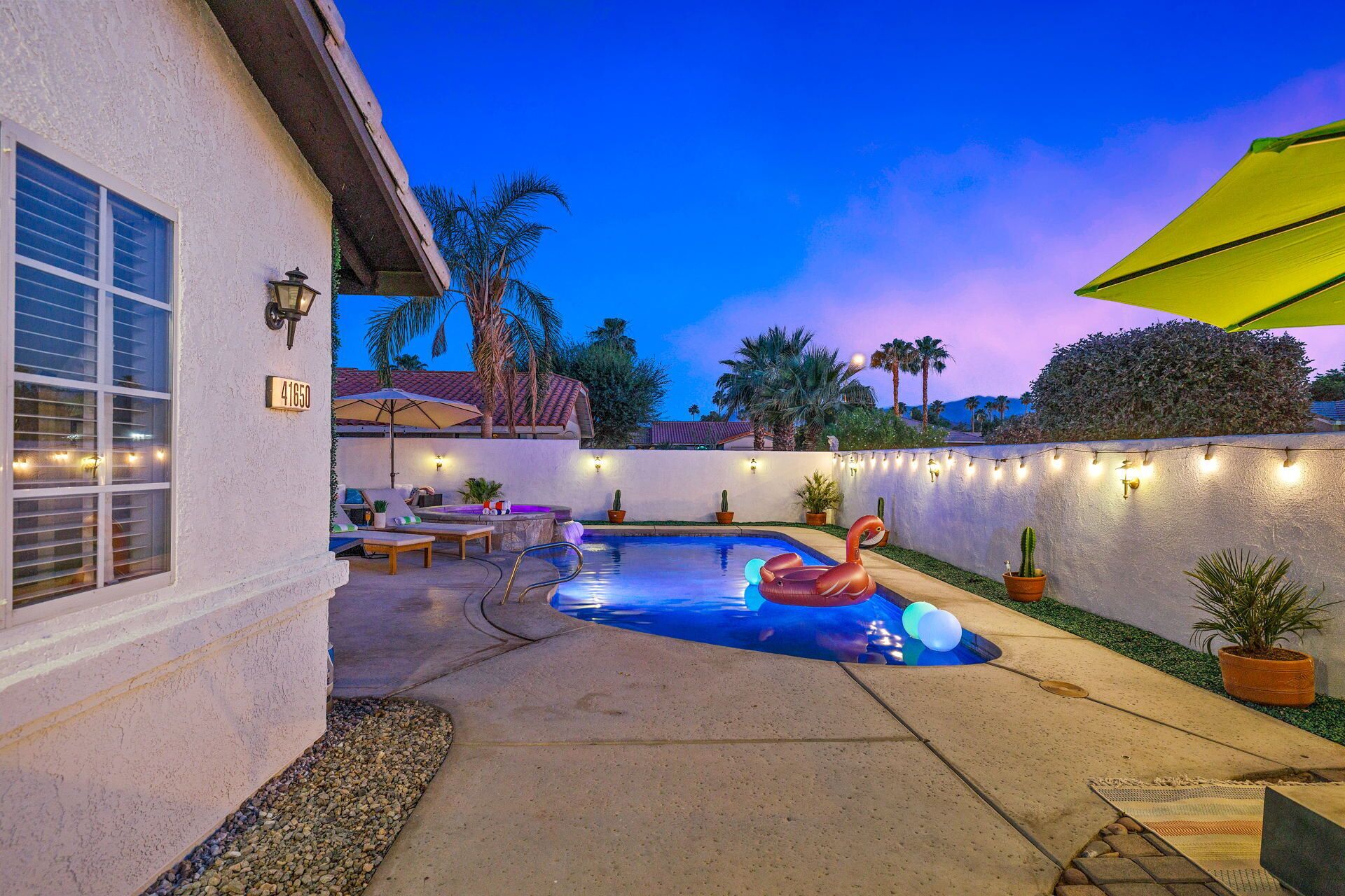 Un patio trasero con piscina, mobiliario exterior e iluminación colgante.