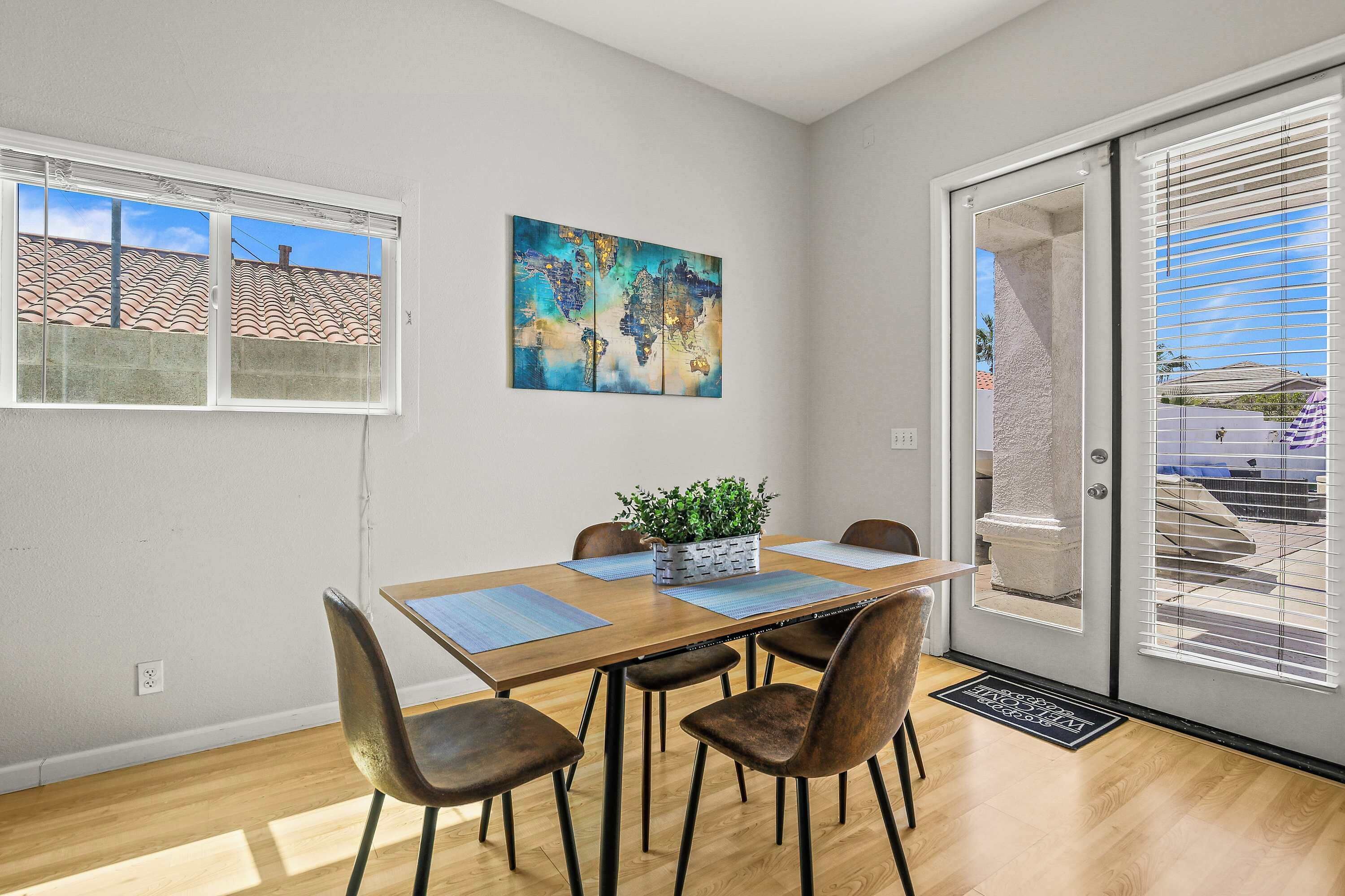 A dining area with a wooden table, four chairs, a world map artwork, and a view of a residential area through a sliding glass door.
