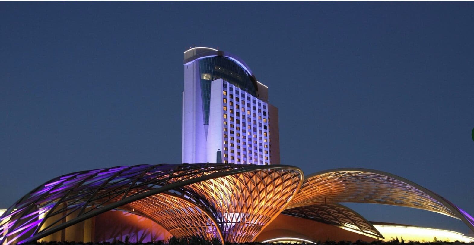 A modern hotel with a distinctive curved roof structure and a tall building with a grid of windows in the background.