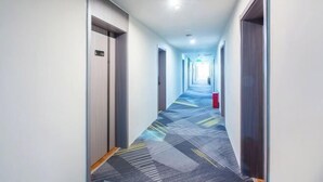 Ein langer, schmaler Hotelgang mit Teppichmuster, Türen an beiden Seiten und einem roten Gegenstand rechts.