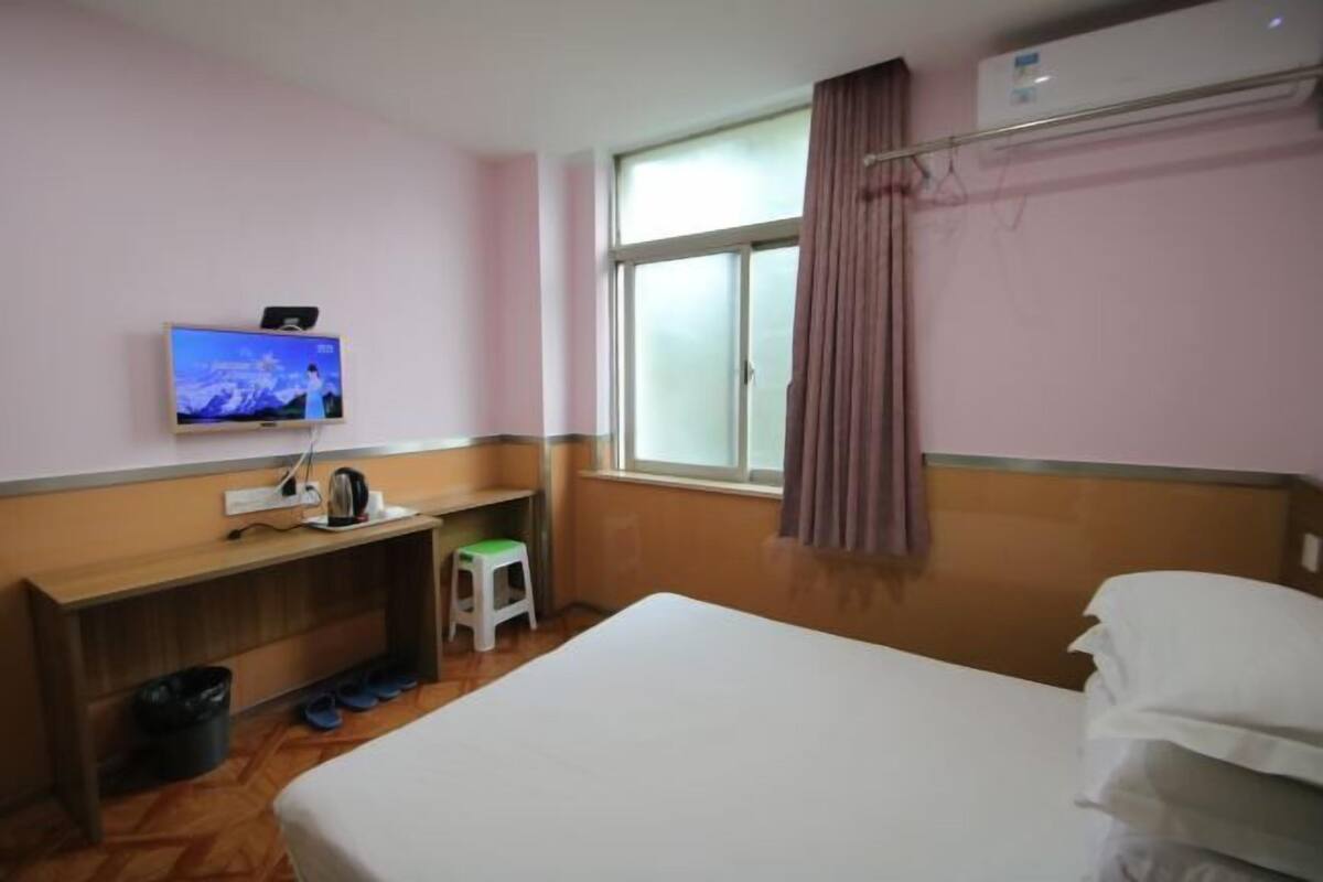 Ein Hotelzimmer mit Bett, Wandfernseher, einem kleinen Hocker, einem Fenster mit Vorhängen und einer Heizung.