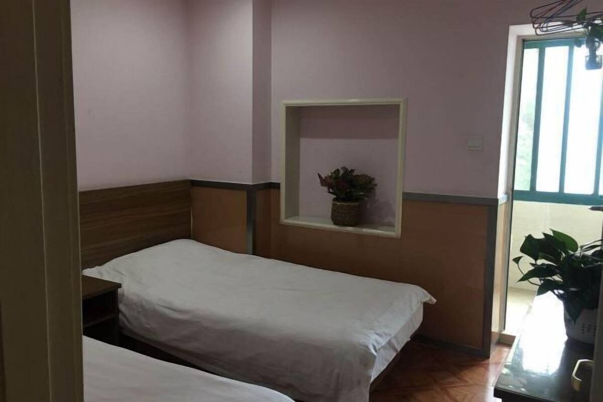 Ein Zimmer mit zwei Einzelbetten, einem kleinen Regal mit einer Pflanze und einem Fenster mit Jalousien.