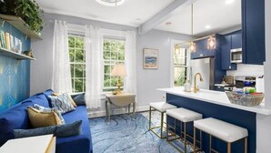 Un salon moderne avec un canapé bleu, une petite table à manger et un coin cuisine doté d’un plan de travail blanc et de meubles de rangement bleus.