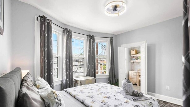 Une chambre à coucher avec un lit, un bureau et une chaise. Il y a des rideaux aux fenêtres et une porte qui donne sur une autre pièce.