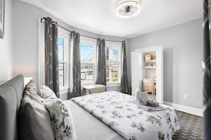 A bedroom with a bed, a desk, and a chair. There are curtains on the windows, and a door leading to another room. - Bright 2BR Retreat • Walk to PATH • NYC Access (Jersey City)