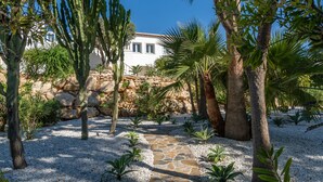 A garden with a stone pathway, cacti, palm trees, and a white building with a tiled roof. - Casa Iris (Javea / Xàbia)