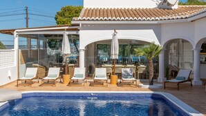 A poolside area with lounge chairs and umbrellas, a clear view of the pool, and a building with a tiled roof and large windows. - Casa Iris (Javea / Xàbia)