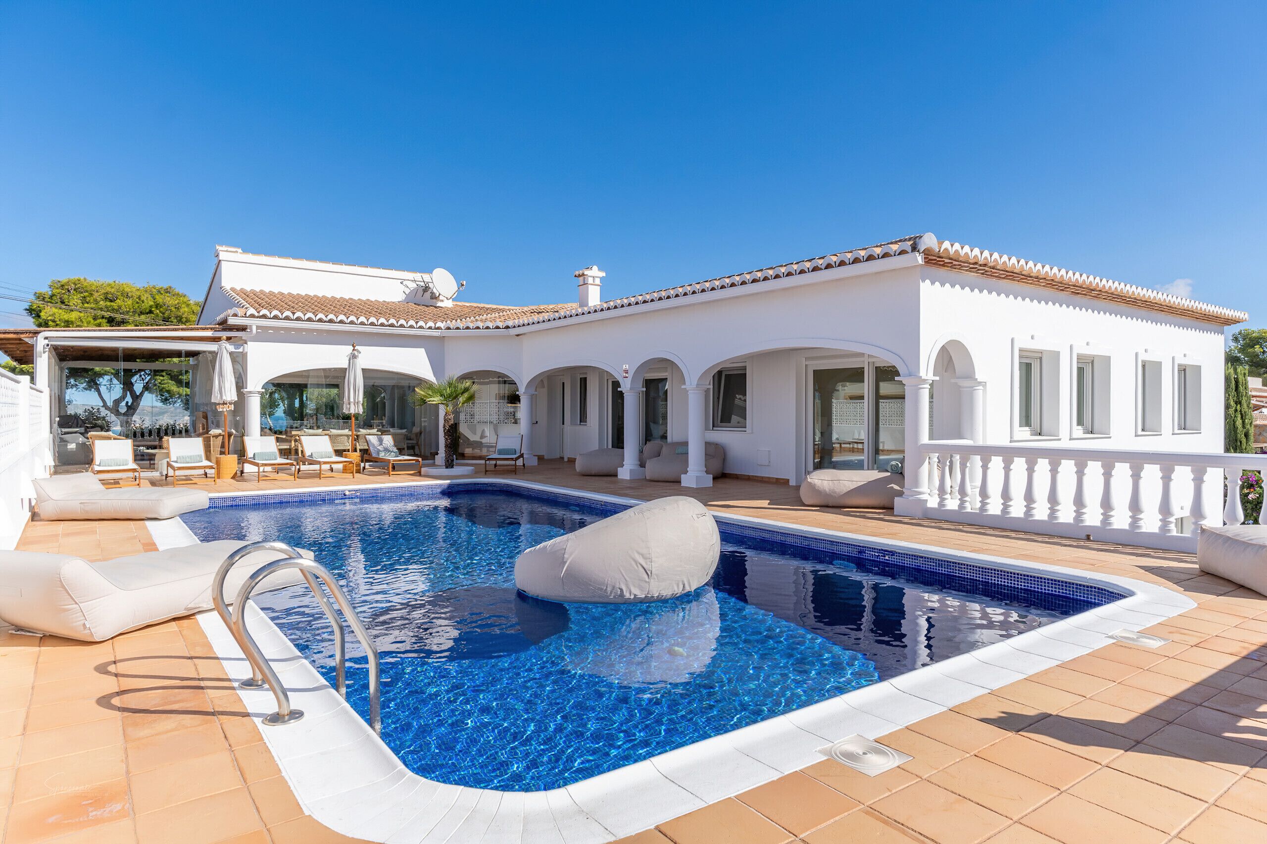 A poolside area with a clear blue pool, white tiled deck, and a white building with arches and columns.